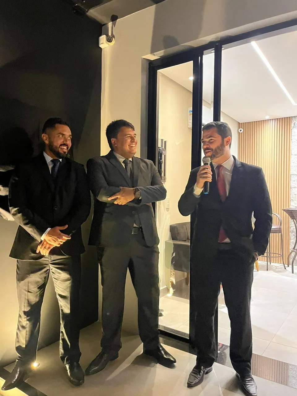Os três sócios na entrada do escritório durante a cerimônia de inauguração; um deles fala ao microfone.