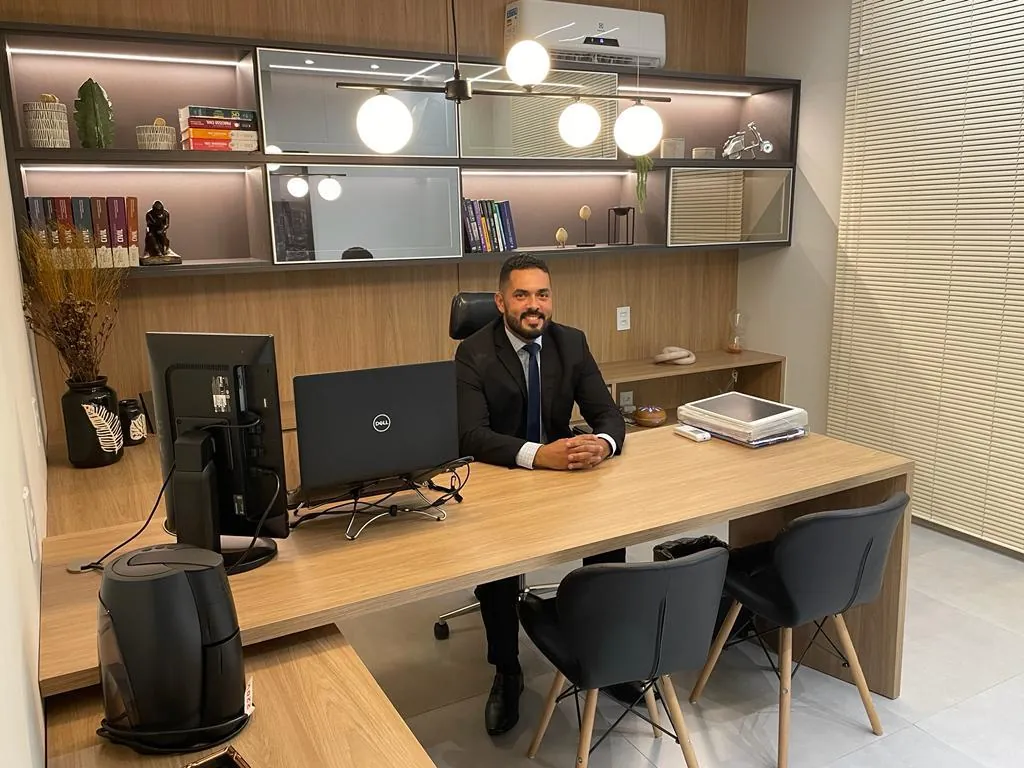 Sala de atendimento com mesa de madeira, cadeiras e estante iluminada; um dos sócios sentado à mesa.
