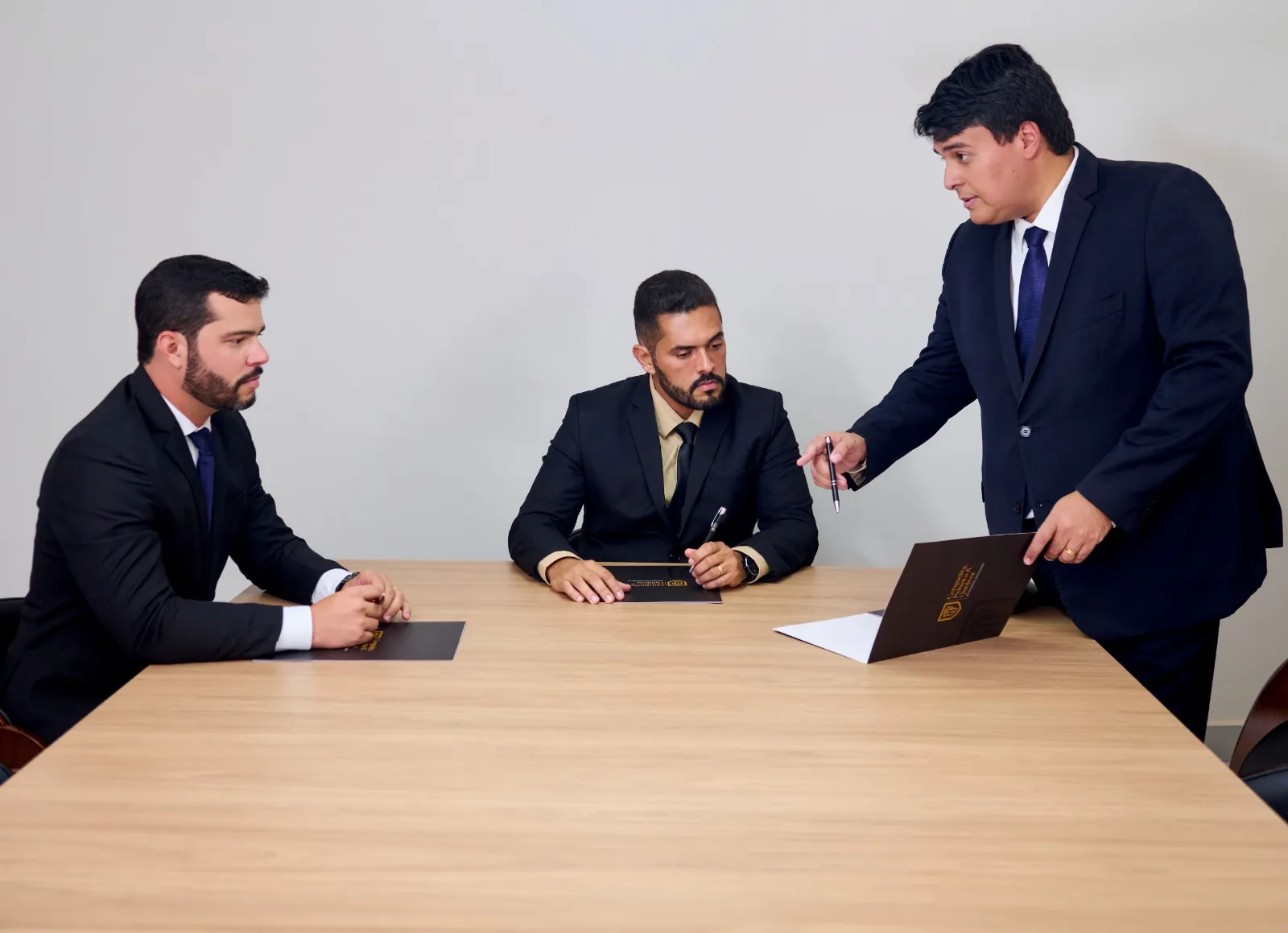 Um dos sócios, em pé, apresenta um documento aos outros dois, sentados à mesa.